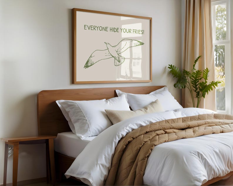 Pode incluir: Uma impress&atilde;o emoldurada com o texto "EVERYONE HIDE YOUR FRIES!" e uma ilustra&ccedil;&atilde;o verde de uma gaivota em voo. A impress&atilde;o est&aacute; acima de uma cama com roupa de cama branca e uma colcha acolchoada castanha. Uma pequena mesa de cabeceira de madeira tamb&eacute;m &eacute; vis&iacute;vel.