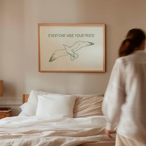 Pode incluir: Uma impress&atilde;o emoldurada com uma ilustra&ccedil;&atilde;o de gaivota e o texto "EVERYONE HIDE YOUR FRIES!". A obra de arte est&aacute; em uma moldura de madeira clara e pendurada acima de uma cama com roupa de cama branca. A gaivota &eacute; verde.