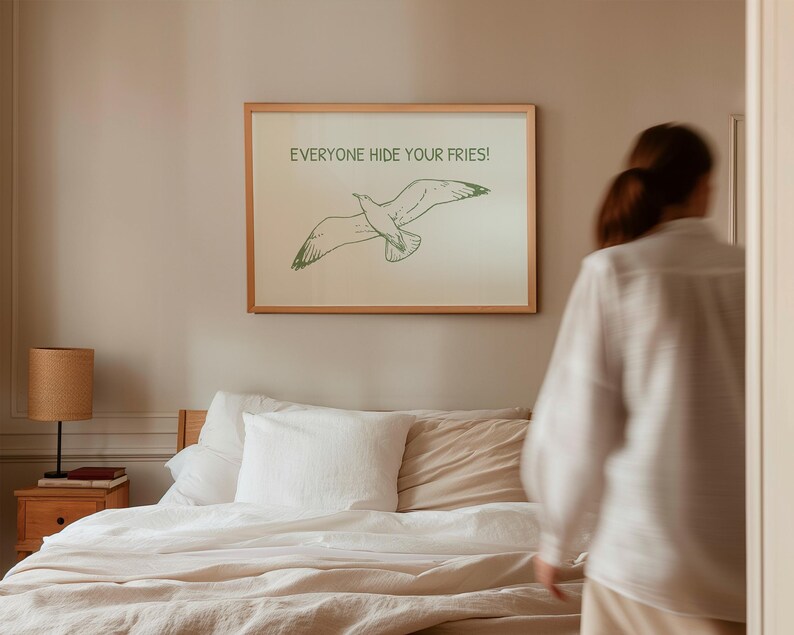 Pode incluir: Uma impress&atilde;o emoldurada com uma ilustra&ccedil;&atilde;o de gaivota e o texto "EVERYONE HIDE YOUR FRIES!". A obra de arte est&aacute; em uma moldura de madeira clara e pendurada acima de uma cama com roupa de cama branca. A gaivota &eacute; verde.
