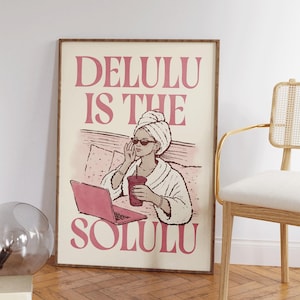 Puede incluir: Una lámina de arte mural rectangular con un dibujo lineal de una persona con una toalla en la cabeza y gafas de sol, sosteniendo una bebida y usando un portátil rosa. El texto DELULU IS THE SOLULU aparece en letras rosas sobre fondo crema.