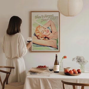 May include: A framed vintage poster with the text "Don't Forget Your SPF" above a retro illustration of a woman sunbathing. The image is displayed in a dining room setting with a table set for a meal.