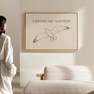 Pode incluir: Impress&atilde;o em tela emoldurada com uma ilustra&ccedil;&atilde;o de gaivota verde e o texto &laquo;EVERYONE HIDE YOUR FRIES!&raquo;. A obra de arte &eacute; emoldurada em uma moldura de madeira e pendurada em uma parede de cor neutra.