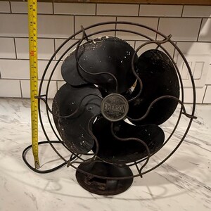Puede incluir: Un ventilador el&eacute;ctrico Emerson antiguo con acabado negro. El ventilador tiene tres aspas y una jaula protectora. El ventilador mide aproximadamente 38 cm de alto. Tiene una base redonda y un emblema met&aacute;lico.