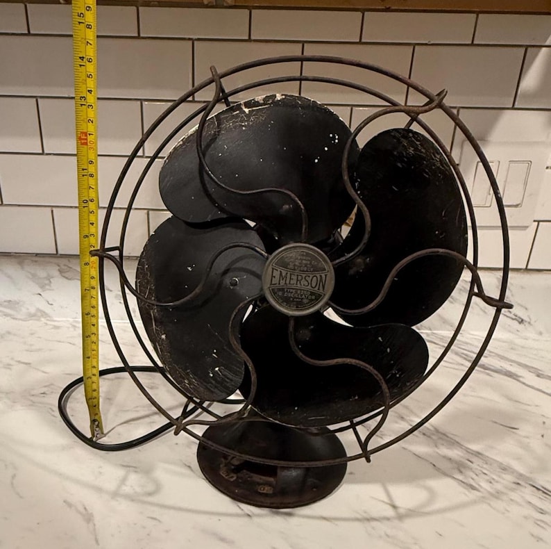 Puede incluir: Un ventilador el&eacute;ctrico Emerson antiguo con acabado negro. El ventilador tiene tres aspas y una jaula protectora. El ventilador mide aproximadamente 38 cm de alto. Tiene una base redonda y un emblema met&aacute;lico.