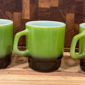 May include: Three vintage green and brown glass mugs. The mugs have a rounded shape with a handle and a dark brown base. The green color fades from light to dark. The mugs are likely from the mid-20th century.