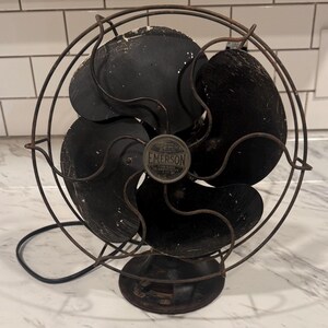 Puede incluir: Un ventilador el&eacute;ctrico Emerson antiguo, con una jaula y aspas negras. El ventilador tiene una base redonda y una jaula met&aacute;lica que rodea las aspas. El logotipo de Emerson es visible en el centro. El ventilador es probablemente vintage y muestra signos de desgaste.