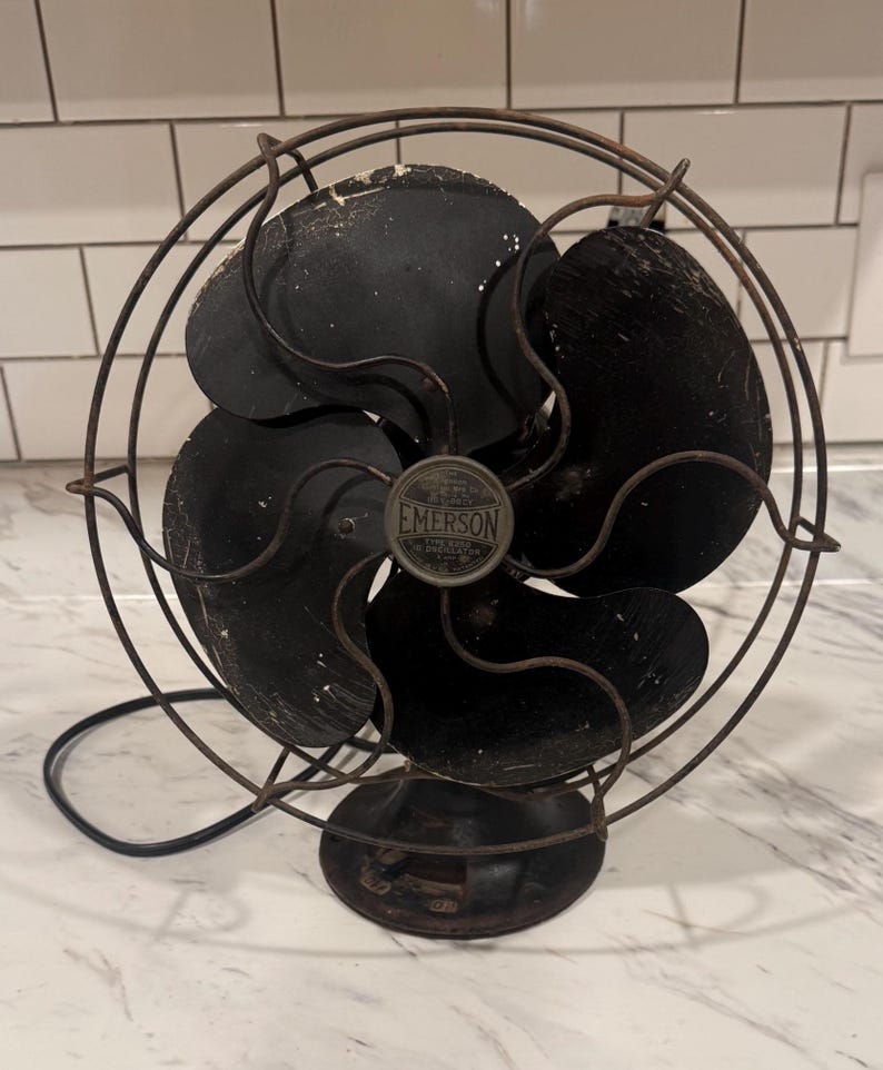 Puede incluir: Un ventilador el&eacute;ctrico Emerson antiguo, con una jaula y aspas negras. El ventilador tiene una base redonda y una jaula met&aacute;lica que rodea las aspas. El logotipo de Emerson es visible en el centro. El ventilador es probablemente vintage y muestra signos de desgaste.