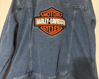 May include: A light blue denim jacket with the Harley-Davidson Cycles logo on the back. The logo is in orange and black. The jacket has a classic design with a button-up front and pockets.