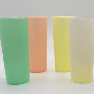 May include: Four tall, pastel-colored drinking glasses. The glasses are in shades of green, peach, yellow, and cream. The glasses are cylindrical and appear to be made of plastic or a similar material. The glasses are empty and are arranged on a white surface.