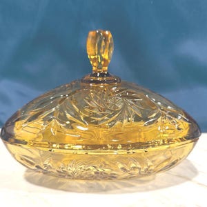 May include: Amber-colored, oval-shaped glass candy dish with a lid. The lid features a decorative finial and a detailed leaf and starburst pattern. The dish is sitting on a white surface with a blue background.