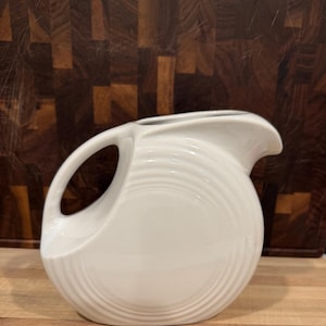 May include: A white ceramic pitcher with a curved handle and a spout. The pitcher has a circular design with concentric ridges. The background is a wooden cutting board.
