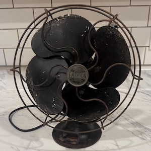 Puede incluir: Ventilador el&eacute;ctrico Emerson vintage con cuatro aspas negras y una jaula protectora de alambre. El ventilador tiene una base redonda y un peque&ntilde;o emblema Emerson en el centro. El ventilador es negro y tiene signos de desgaste.