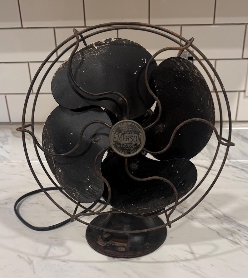 Puede incluir: Ventilador el&eacute;ctrico Emerson vintage con cuatro aspas negras y una jaula protectora de alambre. El ventilador tiene una base redonda y un peque&ntilde;o emblema Emerson en el centro. El ventilador es negro y tiene signos de desgaste.