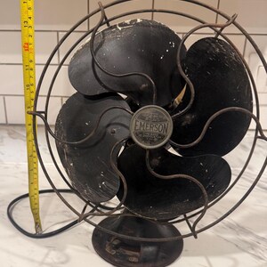 Puede incluir: Ventilador oscilante Emerson vintage con cuatro aspas negras y una jaula protectora de alambre. El ventilador tiene una base negra y una insignia met&aacute;lica que dice "EMERSON". Una cinta m&eacute;trica amarilla est&aacute; al lado del ventilador, midiendo aproximadamente 35 cm.