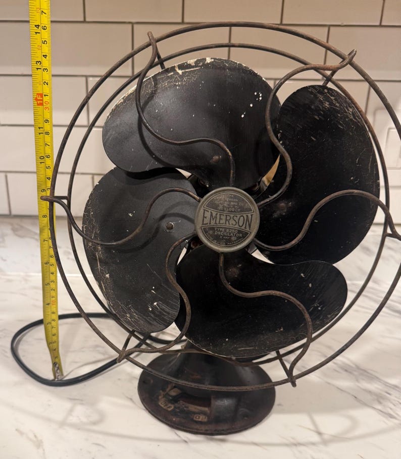 Puede incluir: Ventilador oscilante Emerson vintage con cuatro aspas negras y una jaula protectora de alambre. El ventilador tiene una base negra y una insignia met&aacute;lica que dice "EMERSON". Una cinta m&eacute;trica amarilla est&aacute; al lado del ventilador, midiendo aproximadamente 35 cm.