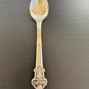 May include: A silver spoon with the word "LUCERNE" engraved on the handle. The spoon's handle features the words "BUCHERER" and "ROLEX" with a rose emblem. The spoon's bowl reflects the surrounding environment.