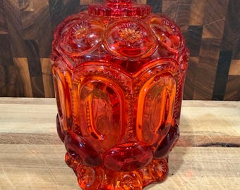 Puede incluir: Un tarro de cristal rojo vibrante con un patrón repetitivo de lunas y estrellas en su superficie. El recipiente cilíndrico incluye una tapa de cristal a juego con un asa de pomo, mostrando un diseño de cristal prensado texturizado.