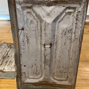 May include: A weathered, rectangular metal panel with a distressed, silver-gray finish. The panel features embossed geometric designs and visible rust along the edges, suggesting age and use. The panel is propped up on a small, ornate gold stand.