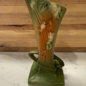 Peut inclure: Un vase en céramique vert et marron avec un haut évasé et deux poignées. Le vase présente un motif floral avec des fleurs blanches et des feuilles vertes. La base est carrée et le vase mesure environ 20 cm de haut.