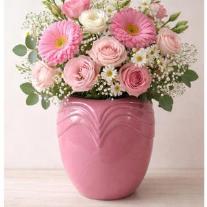 May include: A vibrant floral arrangement in a pink vase. The bouquet features pink gerbera daisies, roses, and small white daisies, with green foliage. The vase is a solid, glossy pink with a heart-shaped design.