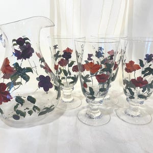 May include: A clear glass pitcher and four matching stemmed glasses. Each piece is decorated with hand-painted red, purple, and blue flowers with green leaves. The pitcher has a large handle and a pouring spout.