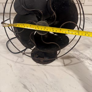 Puede incluir: Un ventilador el&eacute;ctrico negro antiguo con cuatro aspas, encerrado en una jaula de alambre. El ventilador mide aproximadamente 28 cm de di&aacute;metro. Tiene una base redonda y un cable de alimentaci&oacute;n.
