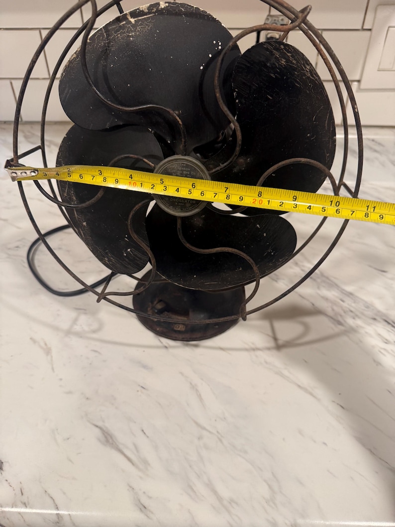 Puede incluir: Un ventilador el&eacute;ctrico negro antiguo con cuatro aspas, encerrado en una jaula de alambre. El ventilador mide aproximadamente 28 cm de di&aacute;metro. Tiene una base redonda y un cable de alimentaci&oacute;n.