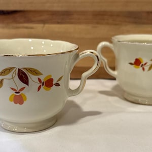 May include: Cream-colored creamer and sugar bowl set. The set features a gold rim and a floral design with red, yellow, and brown leaves and flowers. The creamer has a spout and handle, while the sugar bowl has two handles.