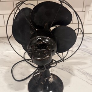 Puede incluir: Un ventilador el&eacute;ctrico negro antiguo con cuatro aspas, una jaula protectora de alambre y una base redonda. El ventilador tiene una est&eacute;tica vintage, con signos de desgaste, lo que sugiere su edad y uso. El dise&ntilde;o del ventilador es un ejemplo cl&aacute;sico de la tecnolog&iacute;a de principios del siglo XX.