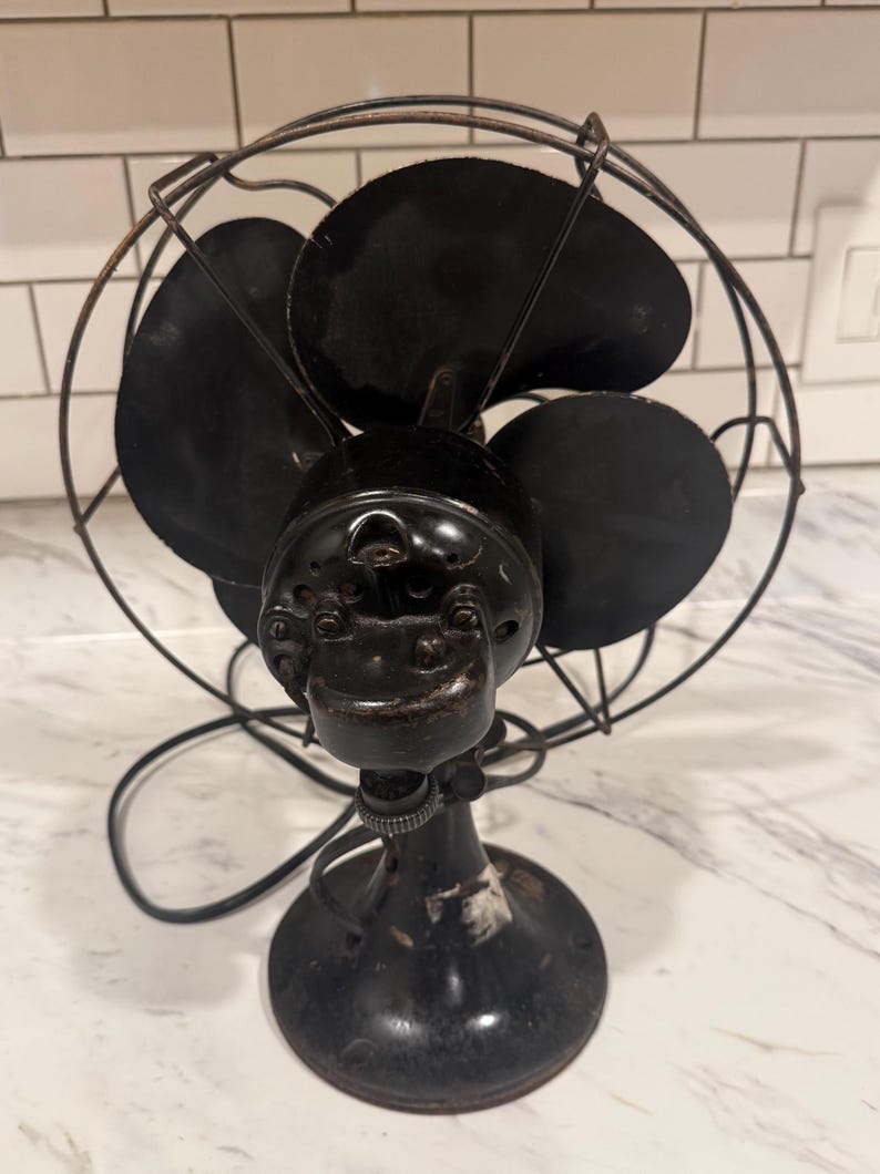 Puede incluir: Un ventilador el&eacute;ctrico negro antiguo con cuatro aspas, una jaula protectora de alambre y una base redonda. El ventilador tiene una est&eacute;tica vintage, con signos de desgaste, lo que sugiere su edad y uso. El dise&ntilde;o del ventilador es un ejemplo cl&aacute;sico de la tecnolog&iacute;a de principios del siglo XX.