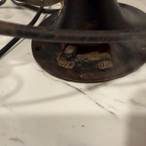 Puede incluir: Primer plano de una base de ventilador el&eacute;ctrico negro antiguo con las palabras "OFF" y "ON" visibles. El ventilador tiene una base circular con un interruptor y una jaula de metal. El ventilador est&aacute; colocado sobre una superficie de m&aacute;rmol blanco.
