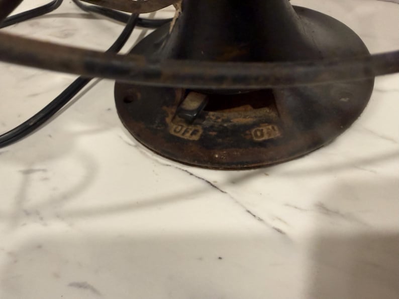 Puede incluir: Primer plano de una base de ventilador el&eacute;ctrico negro antiguo con las palabras "OFF" y "ON" visibles. El ventilador tiene una base circular con un interruptor y una jaula de metal. El ventilador est&aacute; colocado sobre una superficie de m&aacute;rmol blanco.