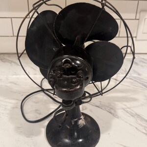 Puede incluir: Un ventilador el&eacute;ctrico negro antiguo con cuatro aspas, encerrado en una jaula de alambre. El ventilador tiene una base redonda y un cable de alimentaci&oacute;n. El ventilador es probablemente vintage.