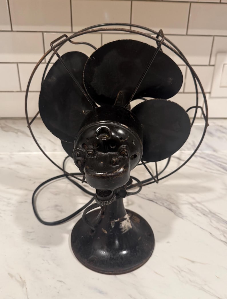 Puede incluir: Un ventilador el&eacute;ctrico negro antiguo con cuatro aspas, encerrado en una jaula de alambre. El ventilador tiene una base redonda y un cable de alimentaci&oacute;n. El ventilador es probablemente vintage.
