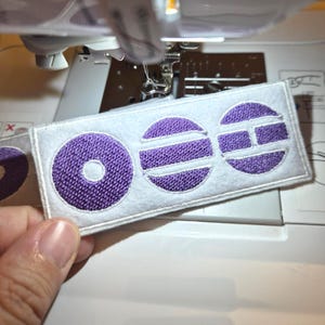 以下が含まれることがあります： 紫色の刺繍された円と文字「OEE」が付いた白いフェルトパッチ。パッチはミシン近くに保持されています。紫色の刺繍は質感のある外観をしています。