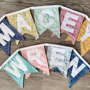 Puede incluir: Una guirnalda de tela con dos filas de banderines que forman el nombre MACEY WREN. Los banderines están hechos de diversas telas estampadas en azul, amarillo, verde, rosa y motivos florales, cada uno con una letra blanca cosida encima.