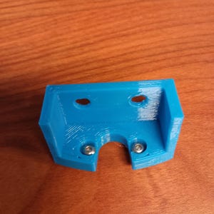 May include: A bright blue, 3D-printed bracket with two screw heads visible. The bracket has a rectangular shape with two holes at the top and a curved opening at the bottom. The item is on a wooden surface.