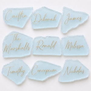 May include: Nine irregular, light blue frosted glass pieces arranged in three rows. Each piece features a unique name or phrase written in elegant gold cursive script, including Caitlin, Deborah, James, The Marshalls, Ronald, Melissa, Timothy, Concepcion, and Nicholas.