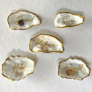 May include: Five oyster shell place cards with gold trim and names: Daniel, Susan, Katherine, Helen, and Harold. The shells are white with a natural texture, and the names are written in elegant gold script.