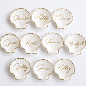 May include: A collection of ten white scallop shells, each featuring a gold-painted rim and a unique name written in gold cursive script. The names displayed on the shells are Shannon, Ally, Olivia, Grace, Karli, Shannon, Bridget, Gabby, Sammi, and Kelley.