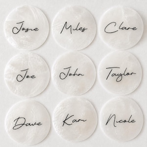 May include: Nine circular white acrylic discs with a marbled texture, each featuring a unique name printed in black cursive script. The names displayed are Josie, Miles, Clare, Joe, John, Taylor, Dave, Kam, and Nicole, arranged in a three-by-three grid.