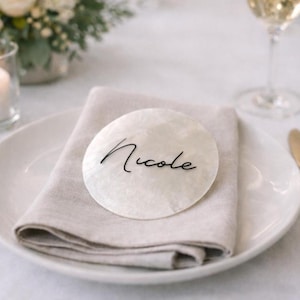 May include: A circular, white place card with the name "Nicole" written in black script. It rests on a folded beige linen napkin, placed on a white plate. The scene suggests a wedding or formal dinner.