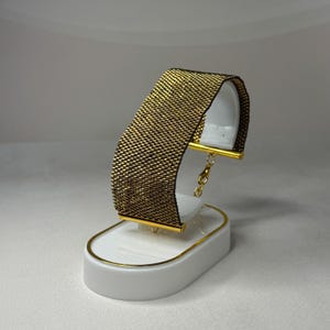 May include: A gold and black beaded bracelet displayed on a white stand with gold accents. The bracelet is a wide band with a woven texture, and the clasp is gold. The stand has a curved shape.