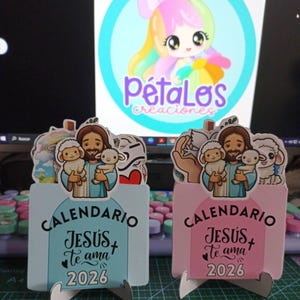 Op de afbeelding: Twee decoratieve kalenders met illustraties van Jezus die lammeren vasthoudt. De ene is lichtblauw, de andere roze. Beide hebben de tekst "CALENDARIO", "JESÚS Te ama" en "2026". Een cartoonlogo is zichtbaar op de achtergrond.