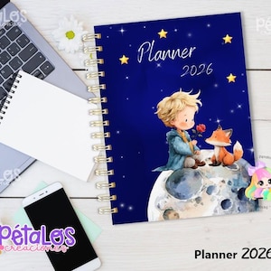 Puede incluir: Una agenda 2026 de espiral con una cubierta azul oscuro que presenta una ilustración de una joven persona de cabello rubio y un zorro sentados sobre la luna, rodeados de estrellas amarillas. El texto dice Planner 2026 en letra cursiva blanca.