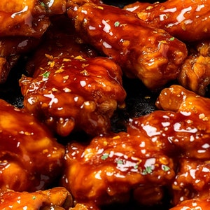 May include: Close-up of a plate of chicken wings covered in a glossy, orange-brown sauce. The wings are arranged in a pile, with some overlapping. The sauce is thick and sticky, and it appears to be made with a combination of soy sauce, honey, and ginger.