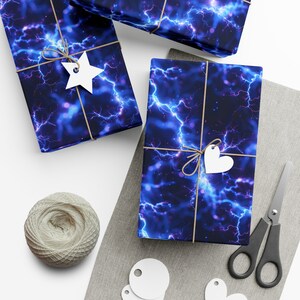 Peut inclure: Trois boîtes cadeaux emballées dans du papier d'emballage à motif d'éclairs bleus et violets. Les boîtes sont attachées avec de la ficelle et décorées d'étoiles et de cœurs en papier blanc.