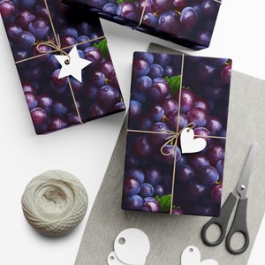 May include: Three gift boxes wrapped in purple and black grape patterned paper with white twine and white heart and star shaped gift tags. The boxes are on a gray fabric surface with white gift tags and a spool of twine.