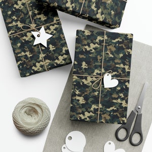 Può includere: Tre scatole regalo avvolte in carta con motivo mimetico, con spago ed etichette di carta bianca. Le etichette hanno la forma di una stella e di un cuore. Le scatole sono su una superficie marrone chiaro.