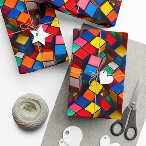 Puede incluir: Tres cajas de regalo envueltas en papel de regalo colorido con un patrón de cubos multicolores. Las cajas están atadas con cuerda y tienen etiquetas de regalo blancas.